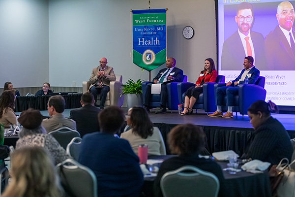 Four health professionals sitting on a stage in chairs while speaking to an audience during a health symposium.
