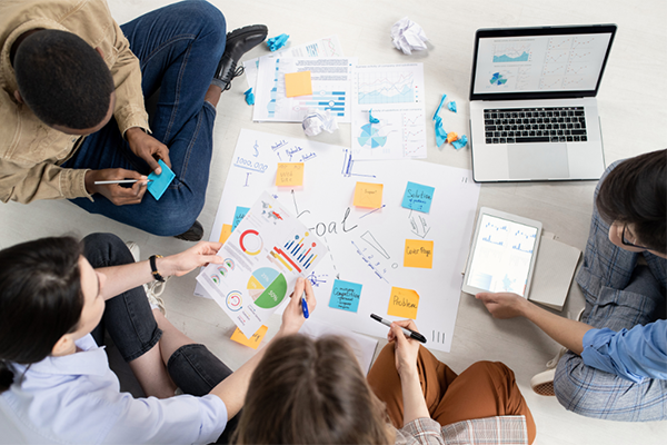 Four design professionals sitting in a circle on the floor, collaborating on a project with notes, papers, and a laptop spread out in front of them.