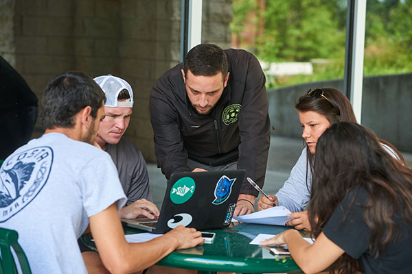 Five students outdoors at a table studying using a laptop and taking notes.