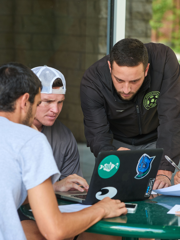 Three students outdoors at a table studying using a laptop and taking notes.