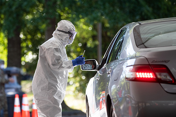 Public health worker wearing a full protection suit standing next to a car outdoors during a drive-through disease testing.
