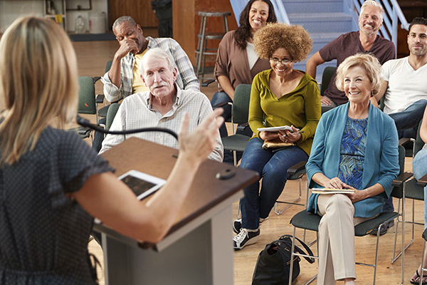Public speaker presenting at a lecturn while a group of people sitting in chairs listen and engage.