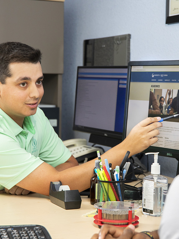 An academic advisor meeting with student in front of a computer at a desk in an office.