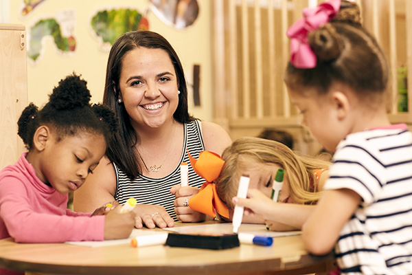 Teacher smiling at camera while coloring on paper with markers with three elementary school students.