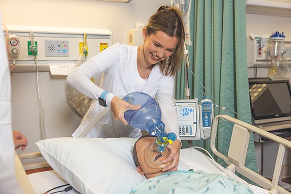 A student using a bag valve mask on a dummy model laying in a hospital bed in a hospital simulation lab.