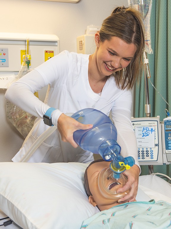A student using a bag valve mask on a dummy model laying in a hospital bed in a hospital simulation lab.