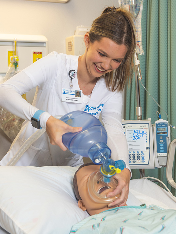 A nursing student using a bag valve mask on a dummy model laying in a bed in the UWF School of Nursing simulation lab.