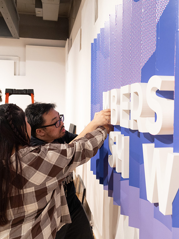 Two students working on a lettered art piece on a wall.