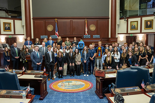 A large group of UWF faculty, staff, administrative leadership, students, student athletes, and UWF mascot Argie the Argonaut pose for a group photo inside the Florida House of Representatives Chamber inside the Florida State Capitol building.