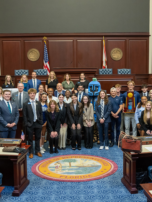 A large group of UWF faculty, staff, administrative leadership, students, student athletes, and UWF mascot Argie the Argonaut pose for a group photo inside the Florida House of Representatives Chamber inside the Florida State Capitol building.
