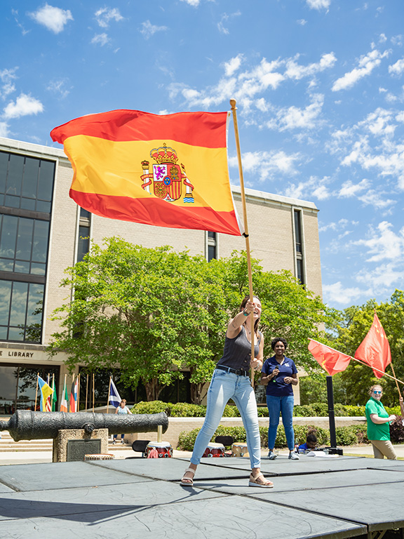 A student carrying a flag pole with the country flag of Spain across a stage during an outdoor event.