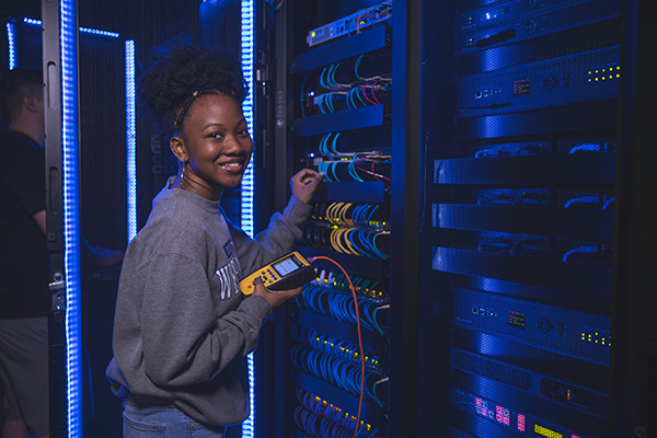 A student smiling at camera while working on cybersecurity related computer equipment.