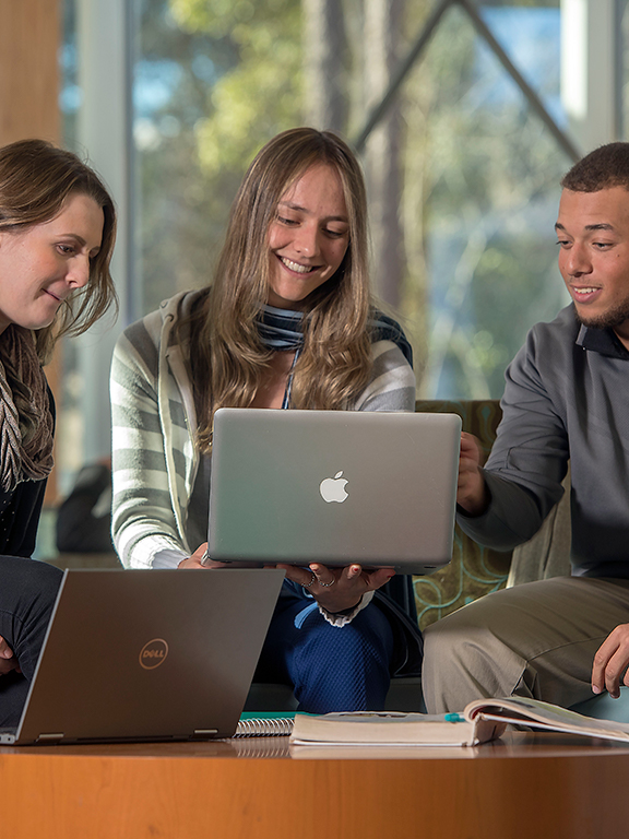Three business students conversing with each other while using a laptop.