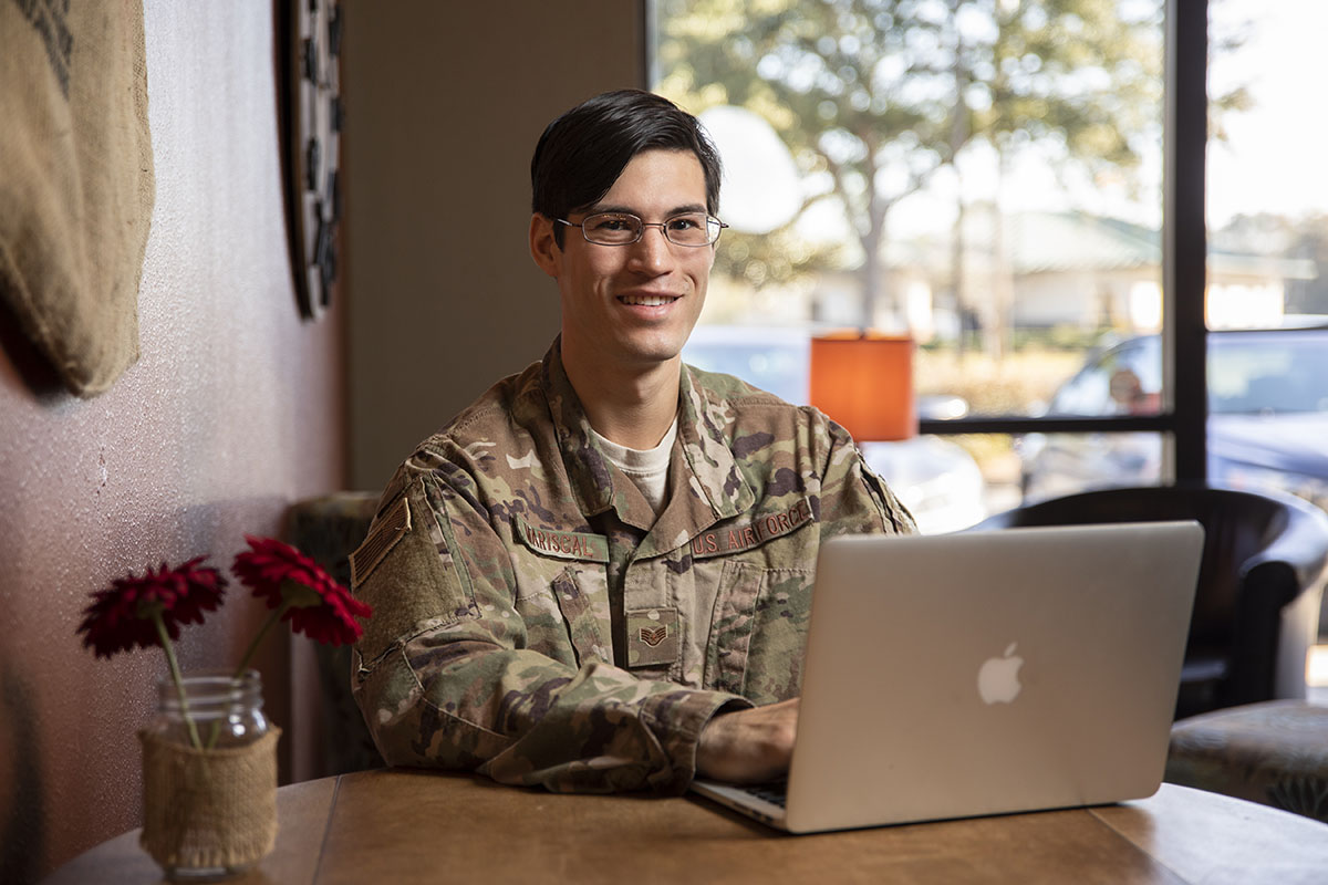 A student in a military uniform smiling at the camera while sitting at a table in a coffee cafe using a laptop.