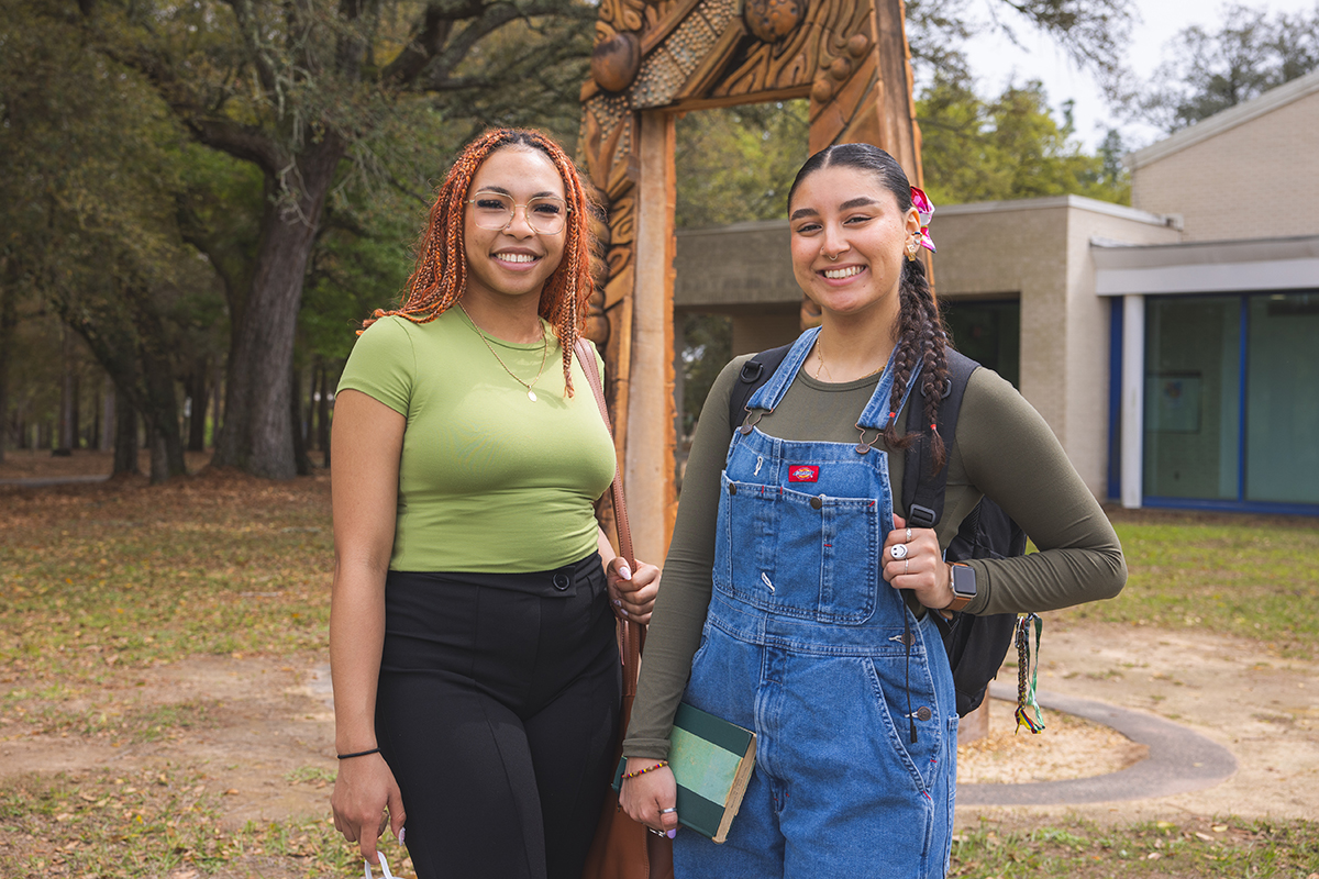 Two students smiling at the camera while standing outdoors in front of an arch art piece in front of a building.