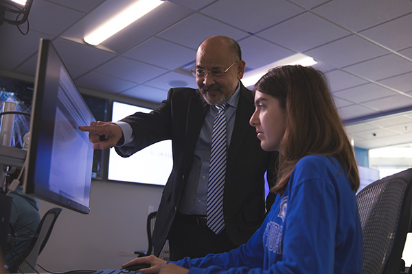 Professor leading a lesson with a student at a computer in a cybersecurity classroom lab.