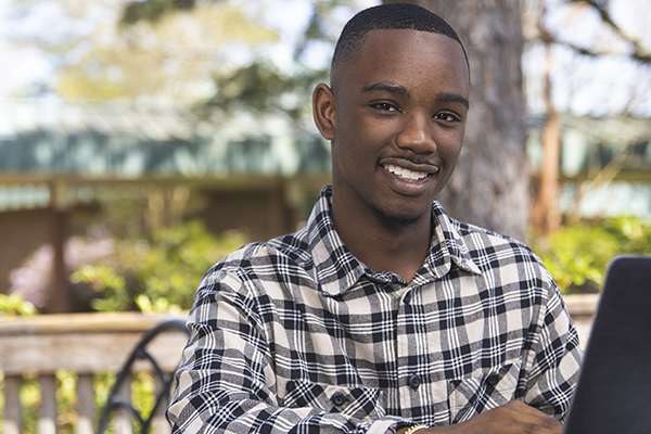 Student smiling at camera while using a laptop outdoors.