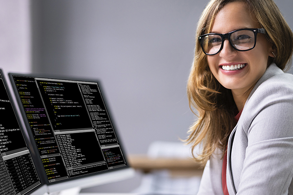 Programmer smiling in front of two computer monitors that display various software code.