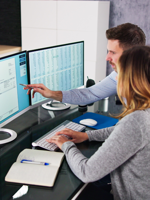 Two financial planners using a computer with two monitors to review an invoice and a financial spreadsheet.