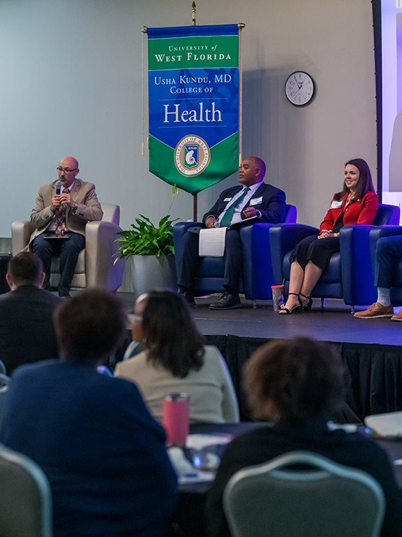Four health professionals sitting on a stage in chairs while speaking to an audience during a health symposium.