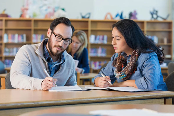 Two students seated at a table in a library collaborating and taking notes