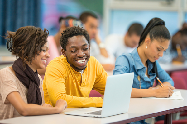 Two students looking at a laptop while seated in a classroom setting with more students in the background taking notes.