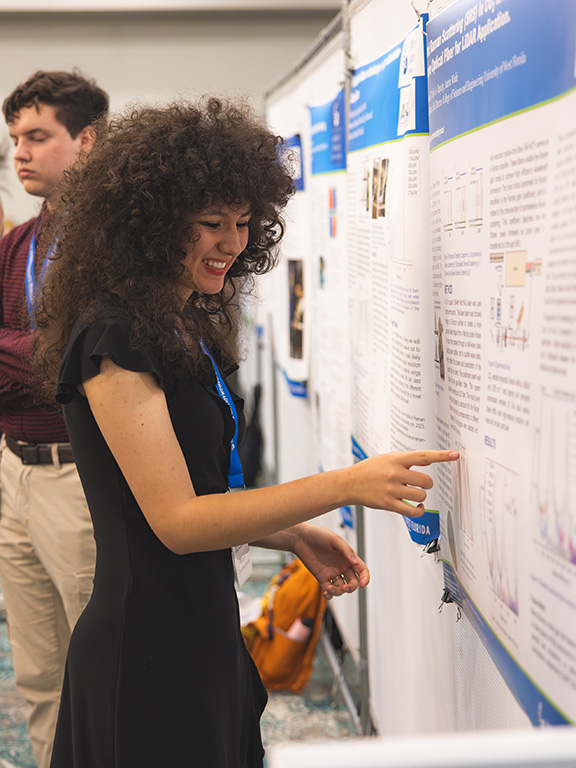 A student pointing to a research project board while presenting to an onlooker during a research symposium.