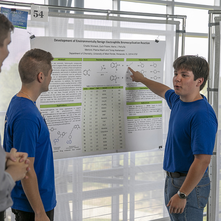 A student presenting a research poster to two onlookers during an indoor research convention.