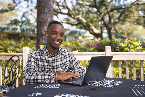 Student smiling at camera while sitting at a table outdoors and using a laptop.
