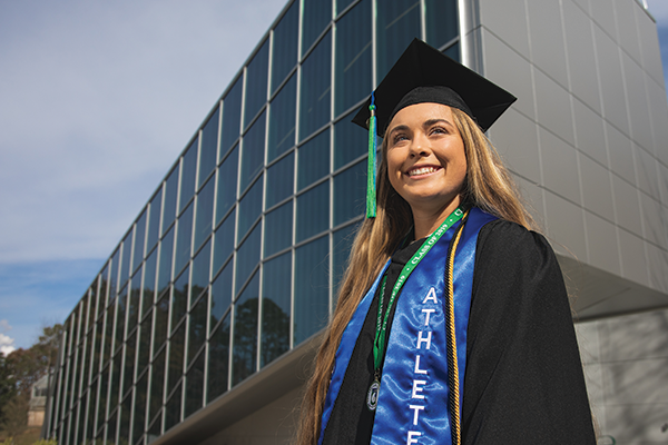 A student in graduation cap and gown attire standing in front of a windowed building wall.