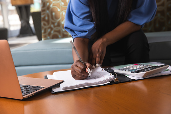 UWF student at a table writing on paper with a pen. A laptop and calculator is on the table to the left and right, respectively, of the student.