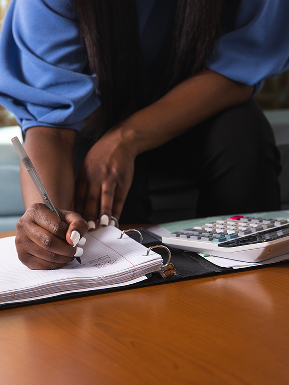 UWF student at a table writing on paper with a pen. A laptop and calculator is on the table to the left and right, respectively, of the student.