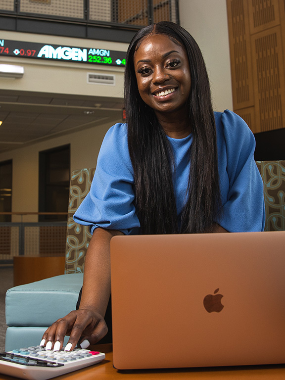 UWF student smiling at camera using a laptop and calculator with a stock market ticker rolling screen in the background.