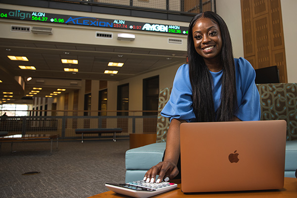 UWF student smiling at camera using a laptop and calculator with a stock market ticker rolling screen in the background.