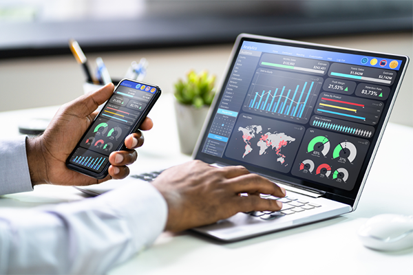 A pair of hands with one hand holding a cellphone and the other hand on the keyboard of a laptop. Both devices display analytical data and charts.