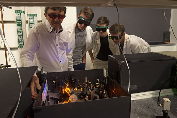physics faculty member and three students wearing goggles examine a physics experiment